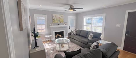 A living room with a fireplace, a ceiling fan, and a large window.