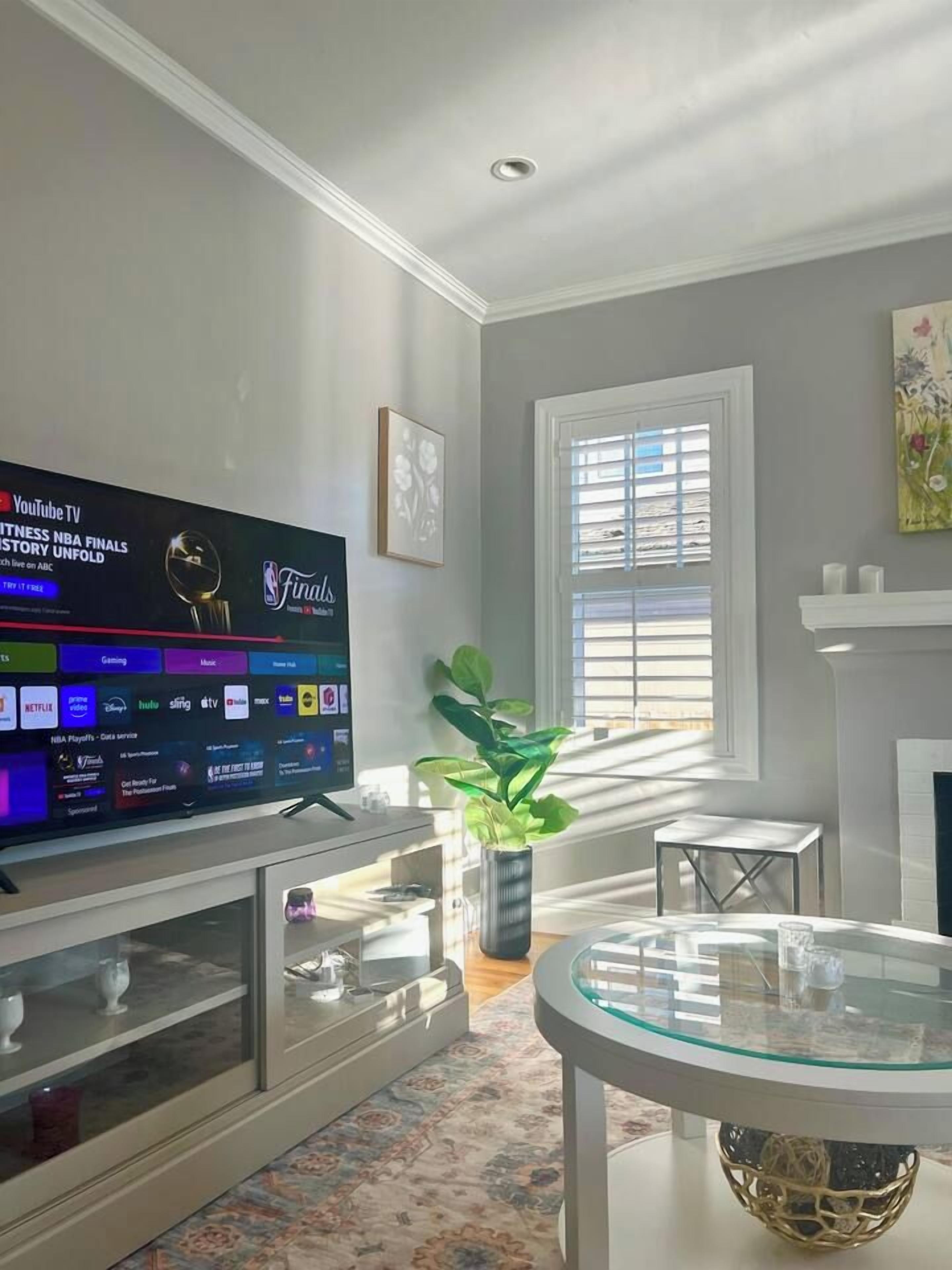 Sala de estar com televisão de tela plana exibindo as finais da NBA, lareira, janela com persianas e um vaso de plantas.