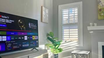 Sala de estar com televisão de tela plana exibindo as finais da NBA, lareira, janela com persianas e um vaso de plantas.