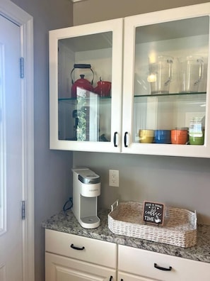 A kitchen with a coffee maker, a basket with a coffee sign, and glass-front cabinets with colorful containers. - Spacious Retreat: Walk to Midtown Near Dining+Fun (Reno)