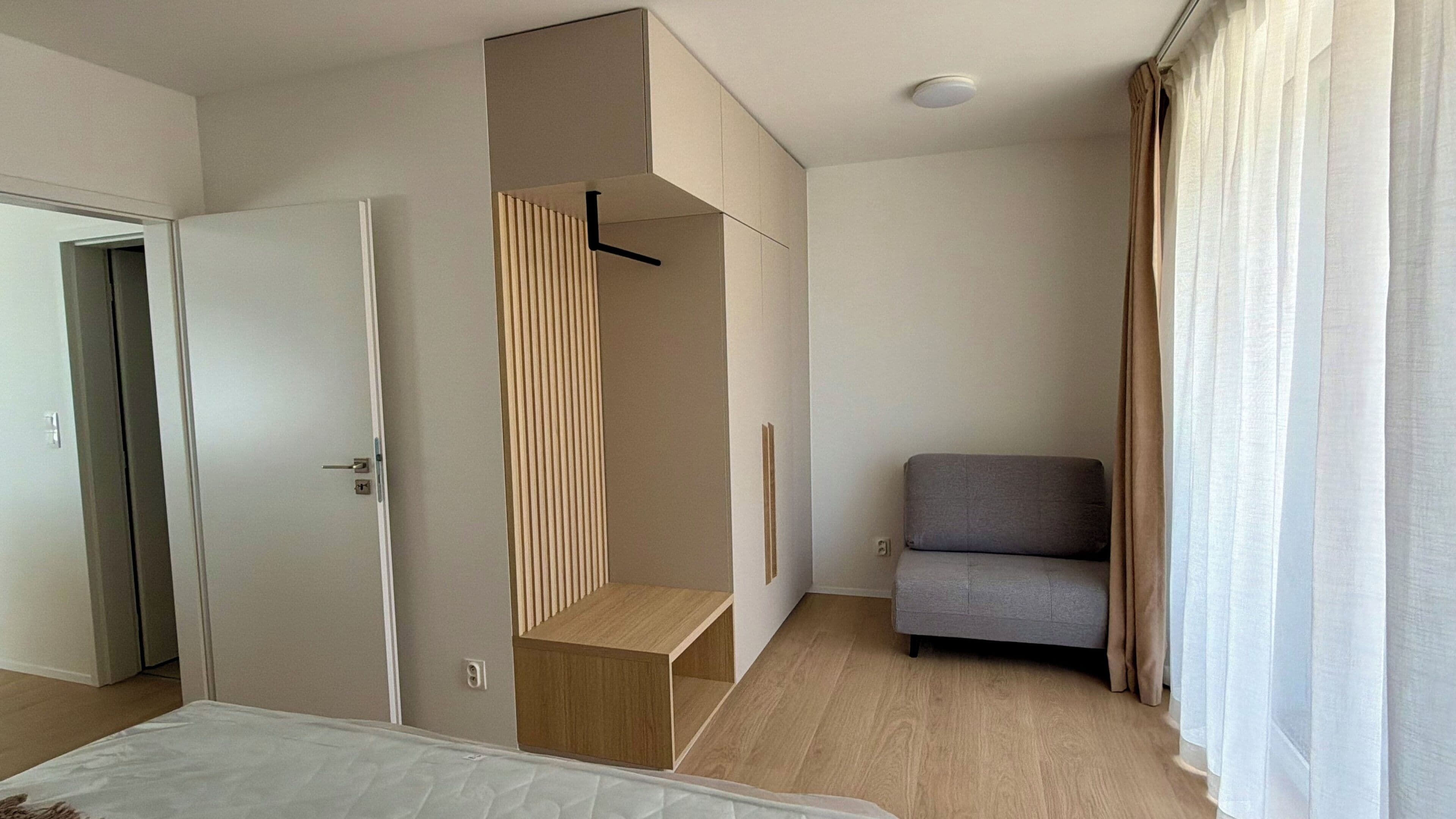 A hotel room with a bed, a seating area with a chair, a wardrobe, and a window with curtains.