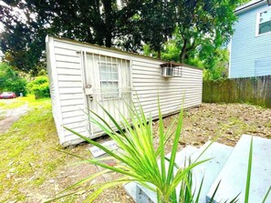 A small, single-story structure with a door and window, surrounded by grass and trees. - Tiny but Mighty Home - 7 mins from DT (Jacksonville)