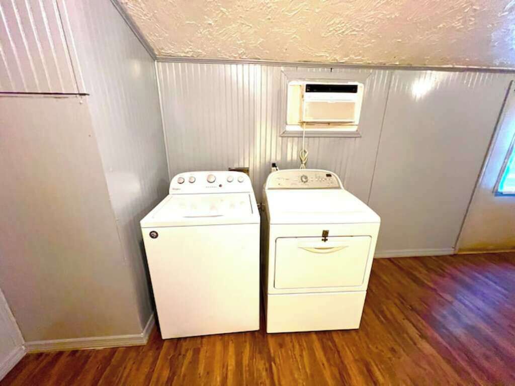 Een wasserette met twee gestapelde wasmachines en drogers, een raam met ventilatie, en een houten vloer.