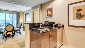 Habitación de hotel con espacio de cocina que incluye un lavabo, estufa y armarios.  Tiene un comedor con mesa y sillas, y obras de arte en las paredes.