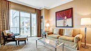 Ein Hotelzimmer mit einer Couch, einem Sessel, einem Hocker, einem Glastisch, einer Stehlampe und einem großen Fenster mit Vorhängen.