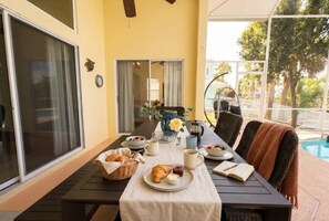 An outdoor dining area with a table set for breakfast, including croissants and coffee, overlooking a pool and greenery. - Camino Del Mar | Canal | Heated Saltwater Pool| Dock +Fenced (Palm Coast)