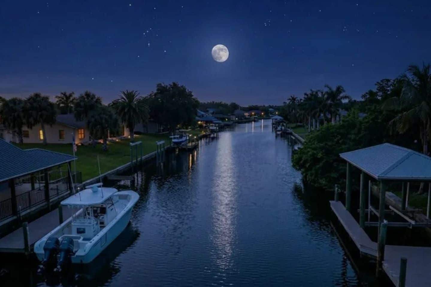 A canal with a boat docked on the left, a covered walkway on the right, and a full moon in the night sky.
