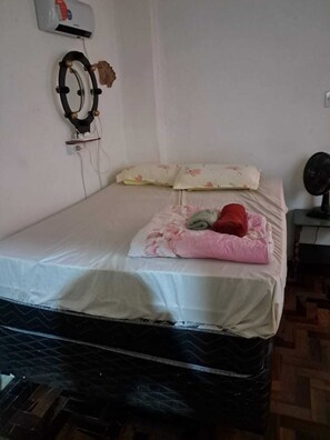 A single bed with a white mattress, a floral pillow, and a red blanket. There is a wall-mounted fan to the right and a power outlet with a plug on the wall. - Apartamento Simples mas Completo Centro Histórico Área Tranquila (Porto Alegre)