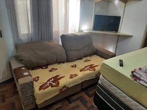 A small, cozy room with a sofa bed, a television mounted on the wall, and a remote control on the bed. - Apartamento Simples mas Completo Centro Histórico Área Tranquila (Porto Alegre)