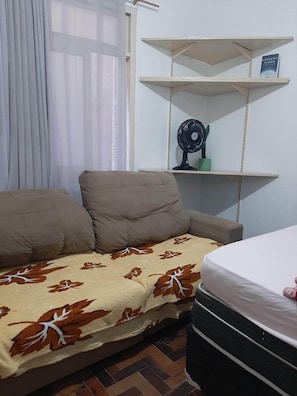 A room with a sofa, a bed, a fan, and shelves. - Apartamento Simples mas Completo Centro Histórico Área Tranquila (Porto Alegre)