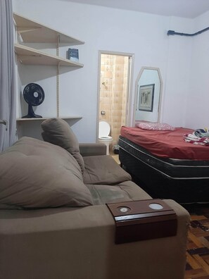 Ein Schlafzimmer mit einem Bett, einem Schlafsofa, einem kleinen Tisch, einem Ventilator, einer Toilette und einem Duschbereich.