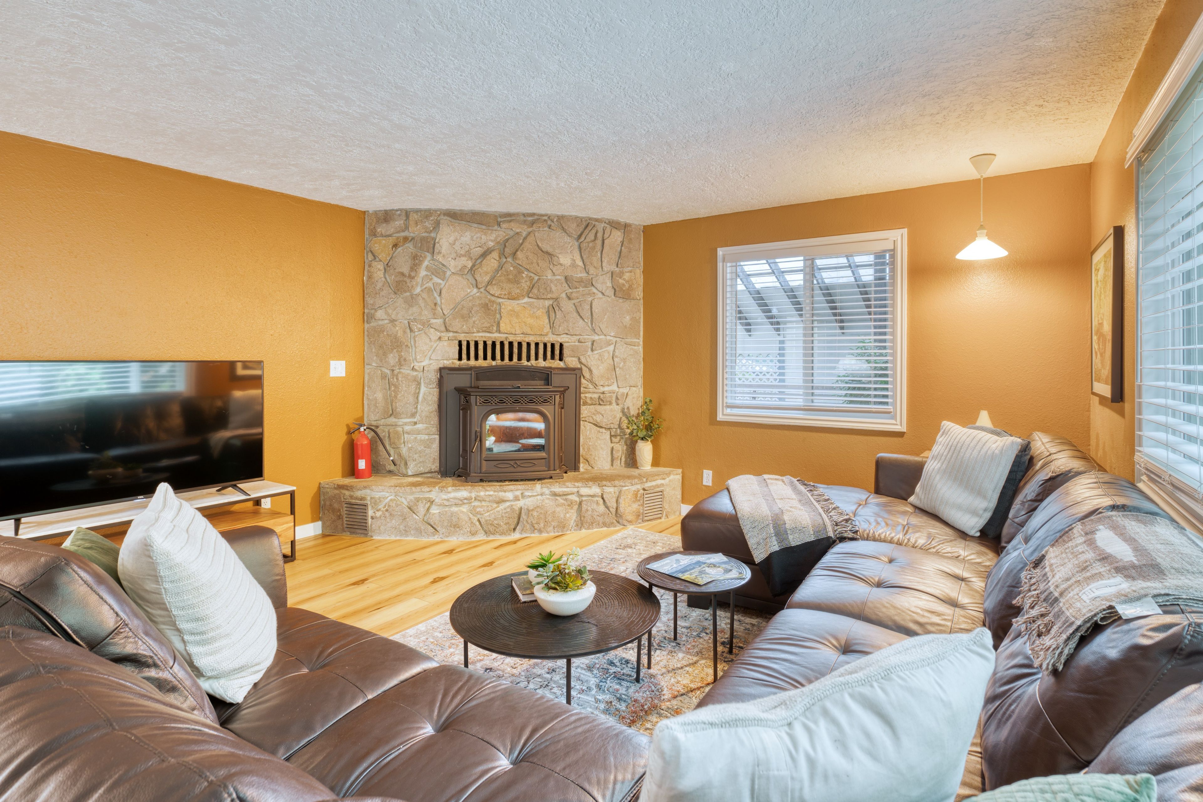 A living room with a stone fireplace, leather sofas, a flat-screen TV, and a large window with blinds.