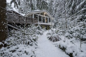 Ein verschneiter Pfad führt zu einer Hütte mit einer Terrasse und großen Fenstern.