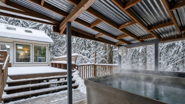 Et overdækket udeområde med jacuzzi, træterrasse og en bygning med glasdør.