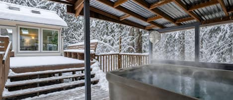 A covered outdoor area with a hot tub, wooden deck, and a building with a glass door.