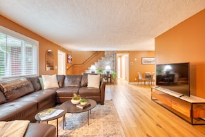A living room with a brown leather sofa, a coffee table with books and plants, a TV on a wooden stand, and a staircase leading to another room.