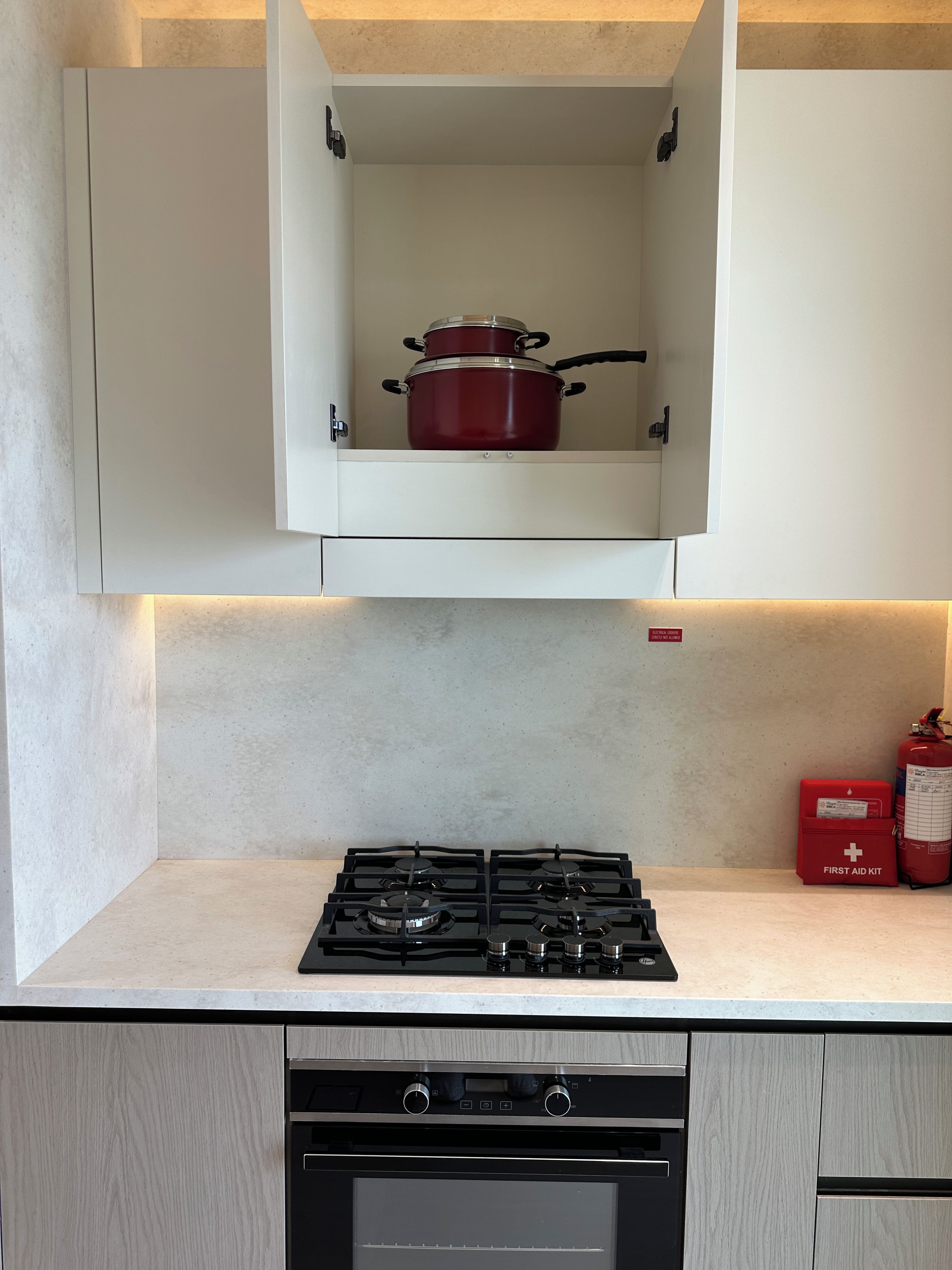 A kitchen with a gas stove, a red pot in a cabinet, and a first aid kit.