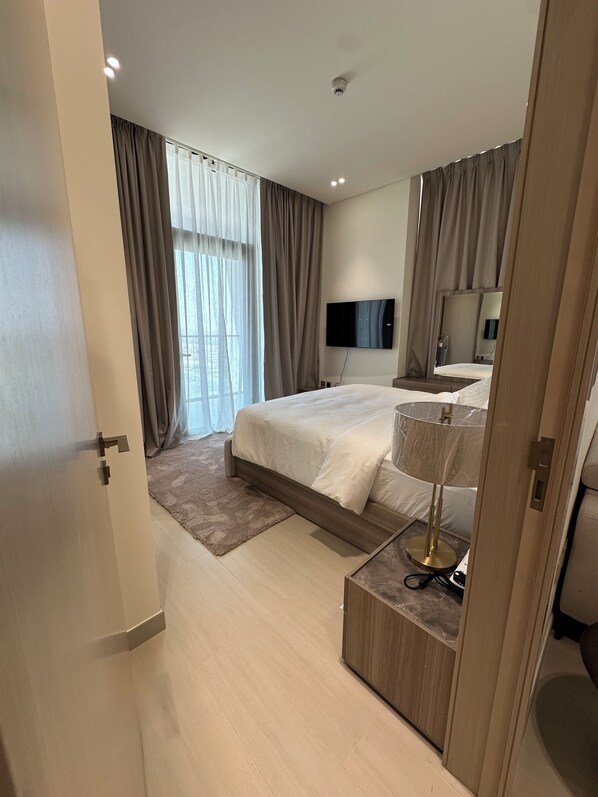 Un dormitorio moderno con una cama grande, un televisor de pantalla plana, una lámpara de noche y vistas al exterior a través de cortinas translúcidas.