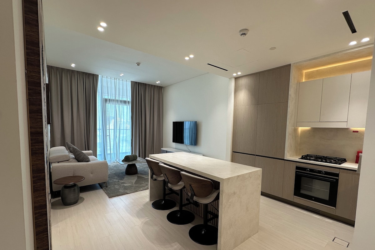 A modern kitchen with a central island, bar stools, a built-in oven, and a flat-screen TV mounted on the wall.