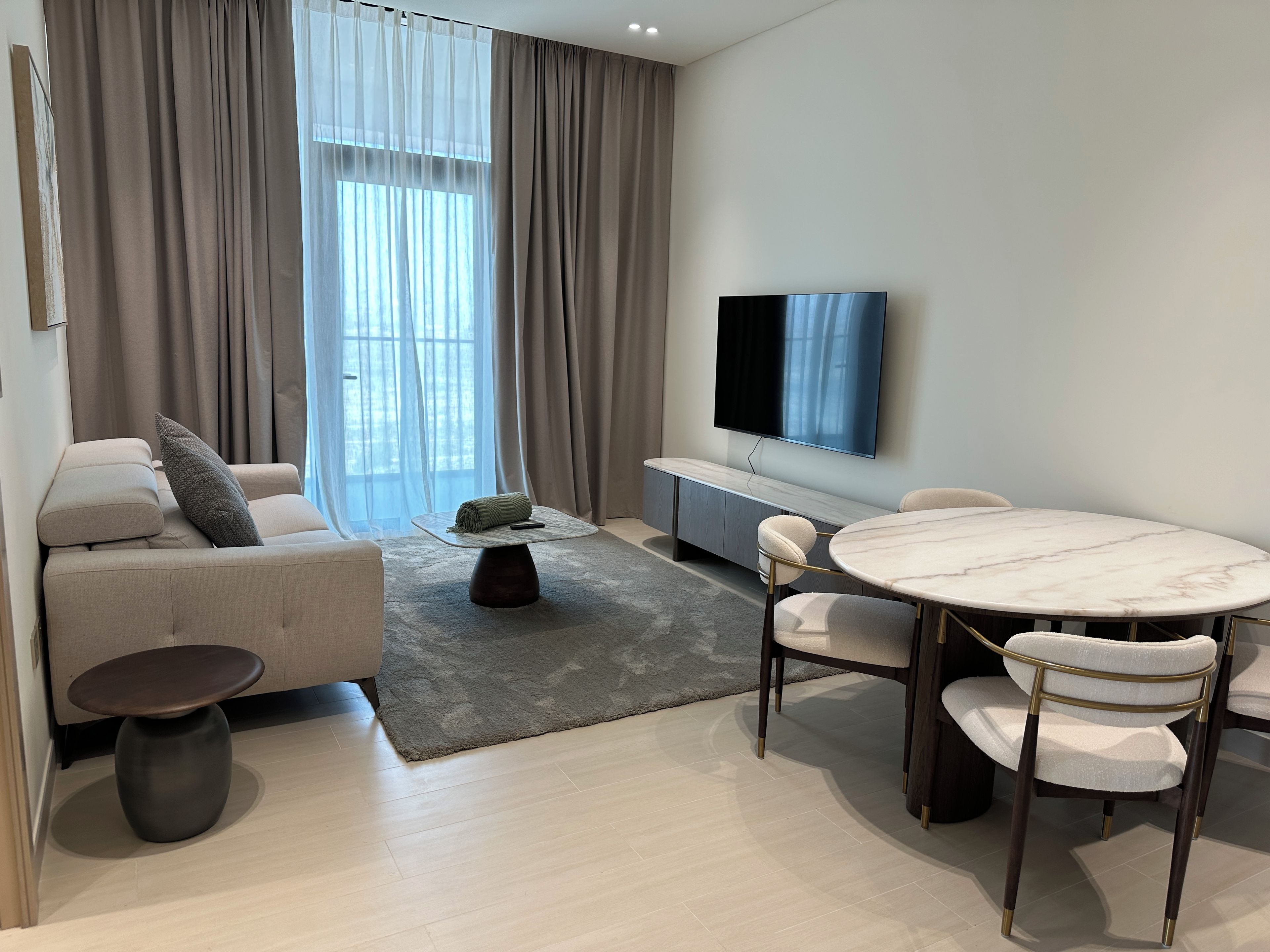 Un salon moderne comprenant un canapé, une table basse, une table à manger avec des chaises, un téléviseur à écran plat et une grande fenêtre avec des rideaux.