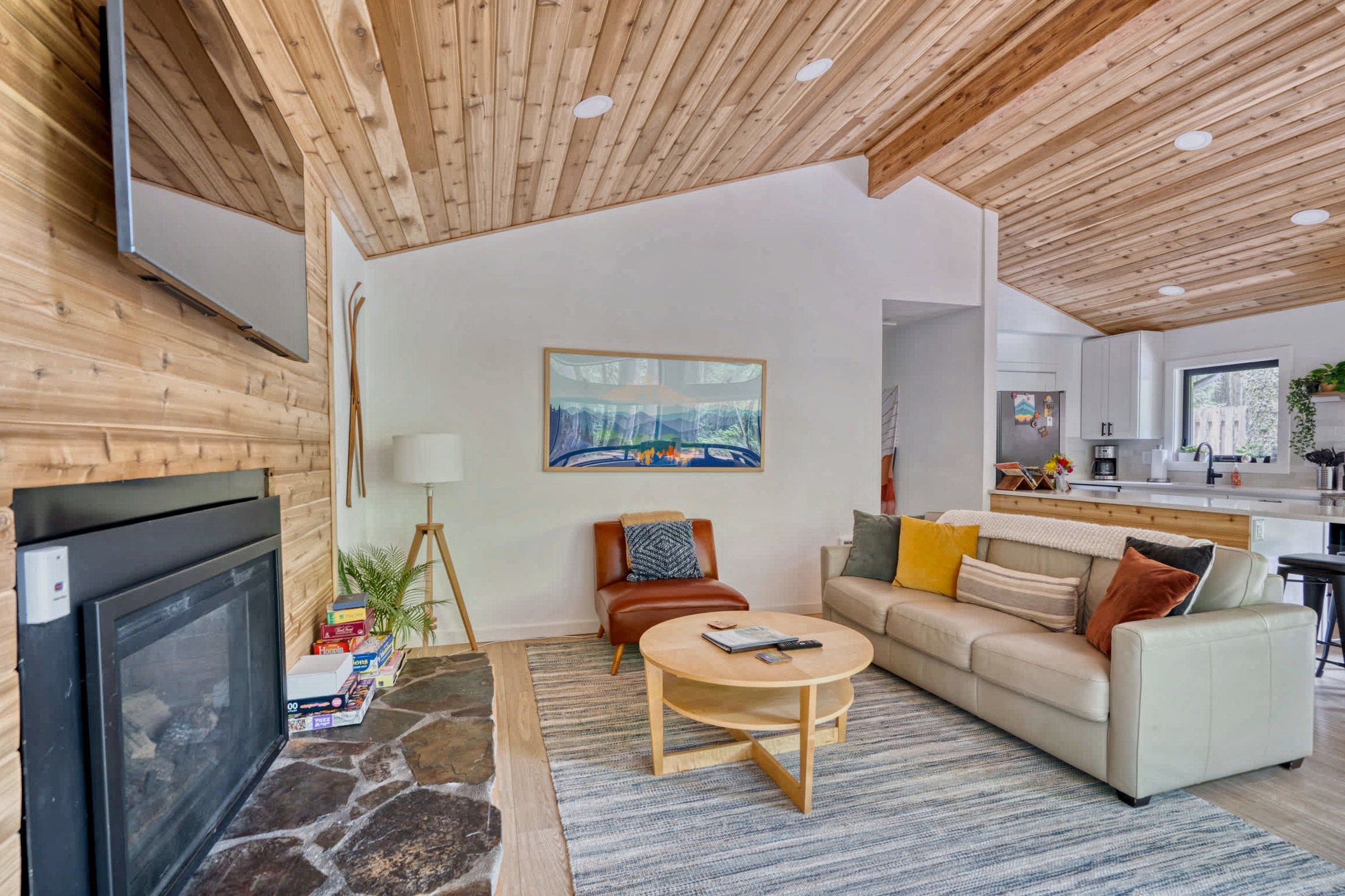 Una sala de estar moderna con chimenea, televisor, un cuadro y una cocina al fondo.