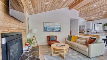 Una sala de estar moderna con chimenea, televisor, un cuadro y una cocina al fondo.