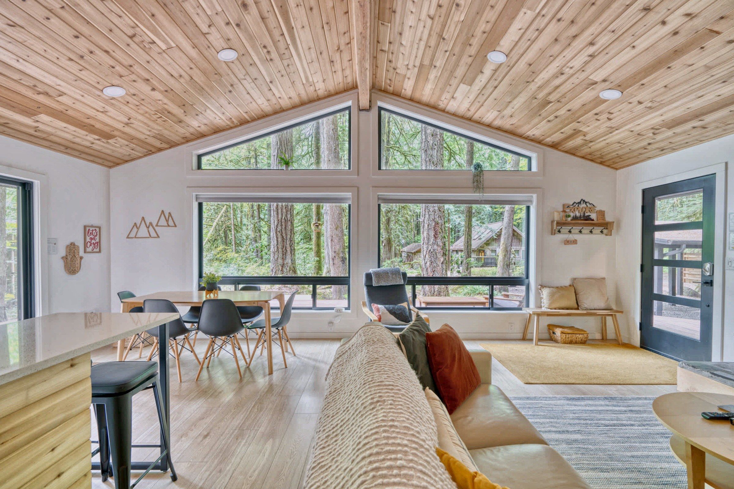 Una habitación amplia con techo alto, ventanas grandes, zona de comedor y espacio de estar con vista al bosque.