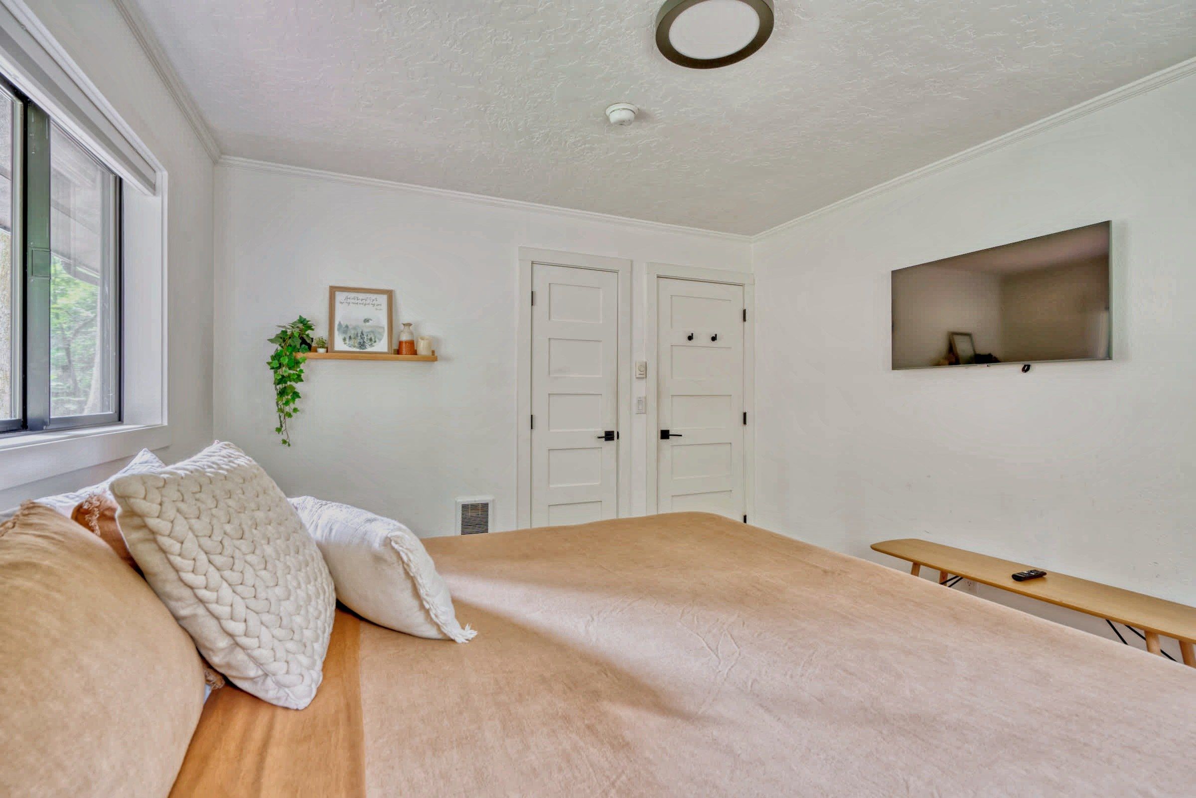 A bedroom with a bed, a bench, a window, a door, and a wall-mounted shelf with a plant and decorative items.