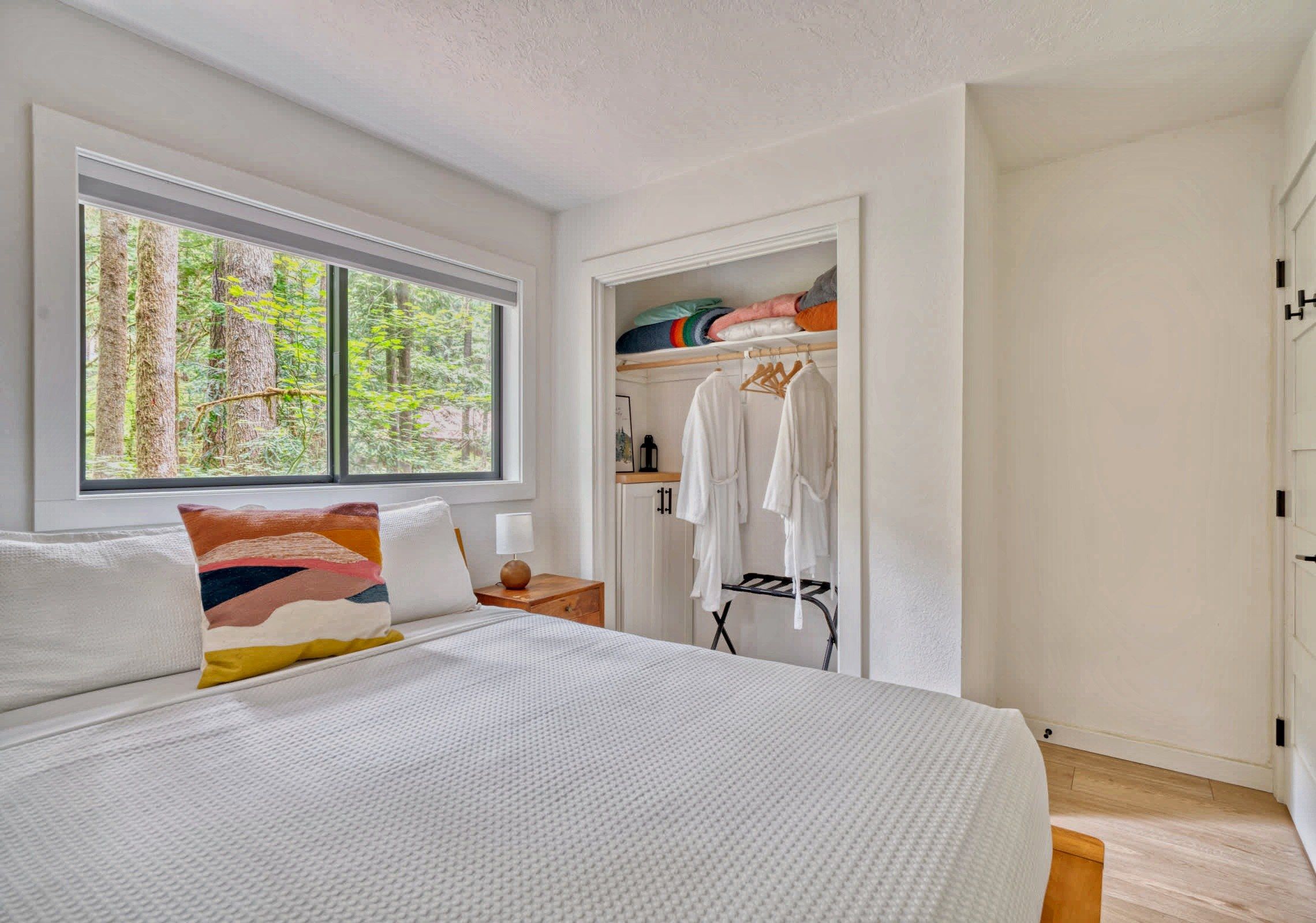 Un dormitorio con una cama grande, una ventana con vista a un bosque, un armario con ropa colgada y una mesita de noche con una lámpara.