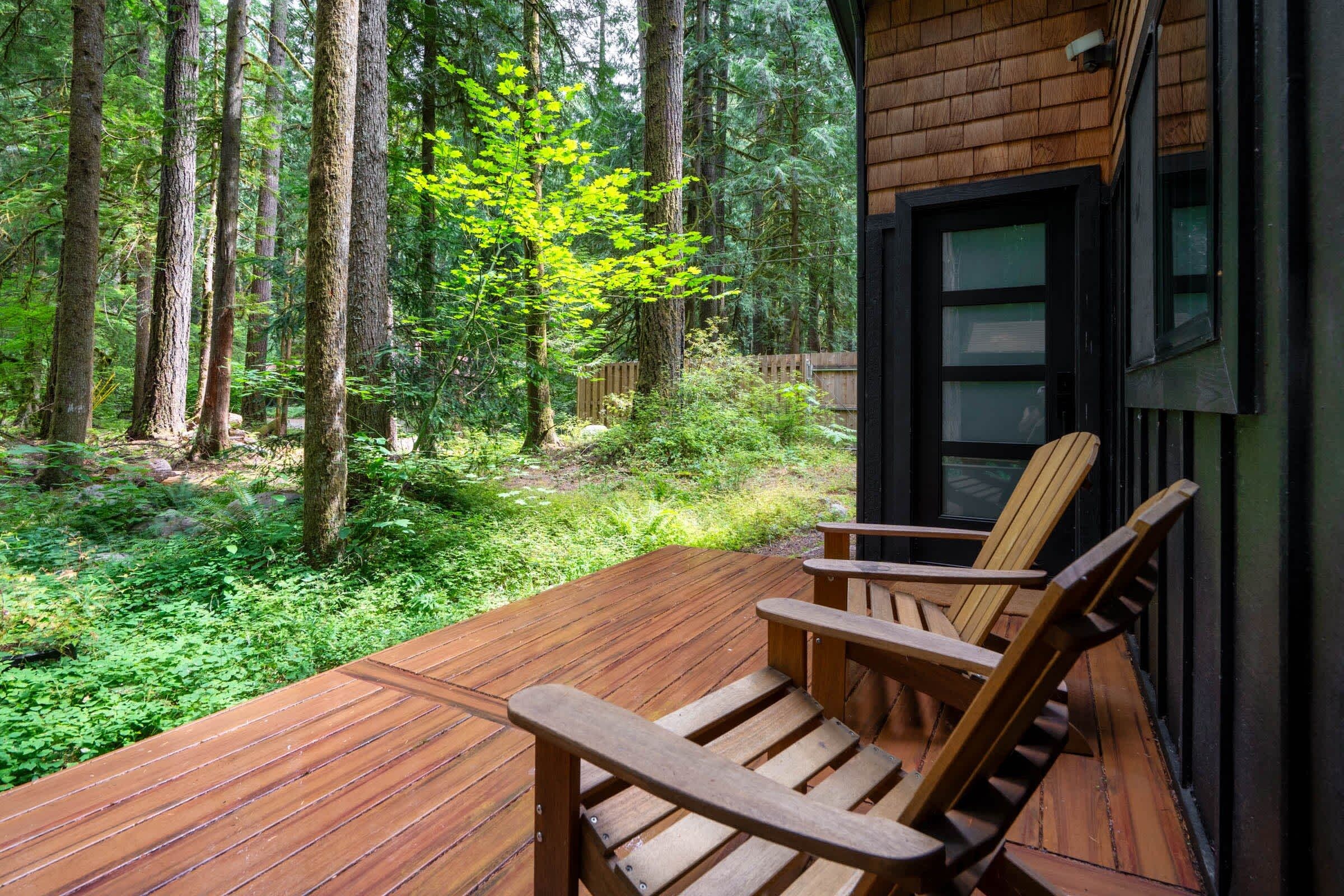 A wooden deck with two chairs, surrounded by a forested area with tall trees and a wooden fence.