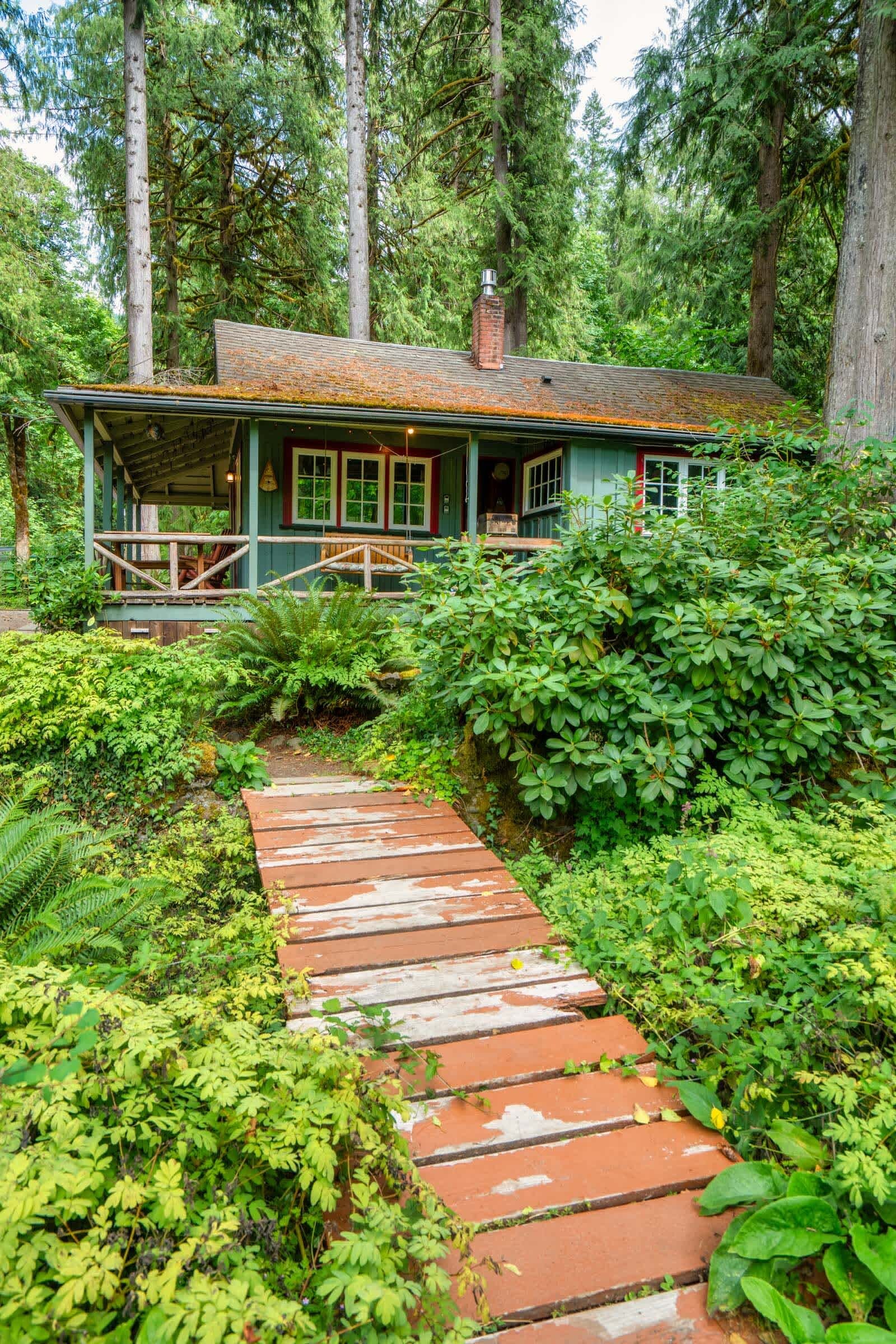 A rustic cabin with a wooden deck leading to a porch, surrounded by dense forest.