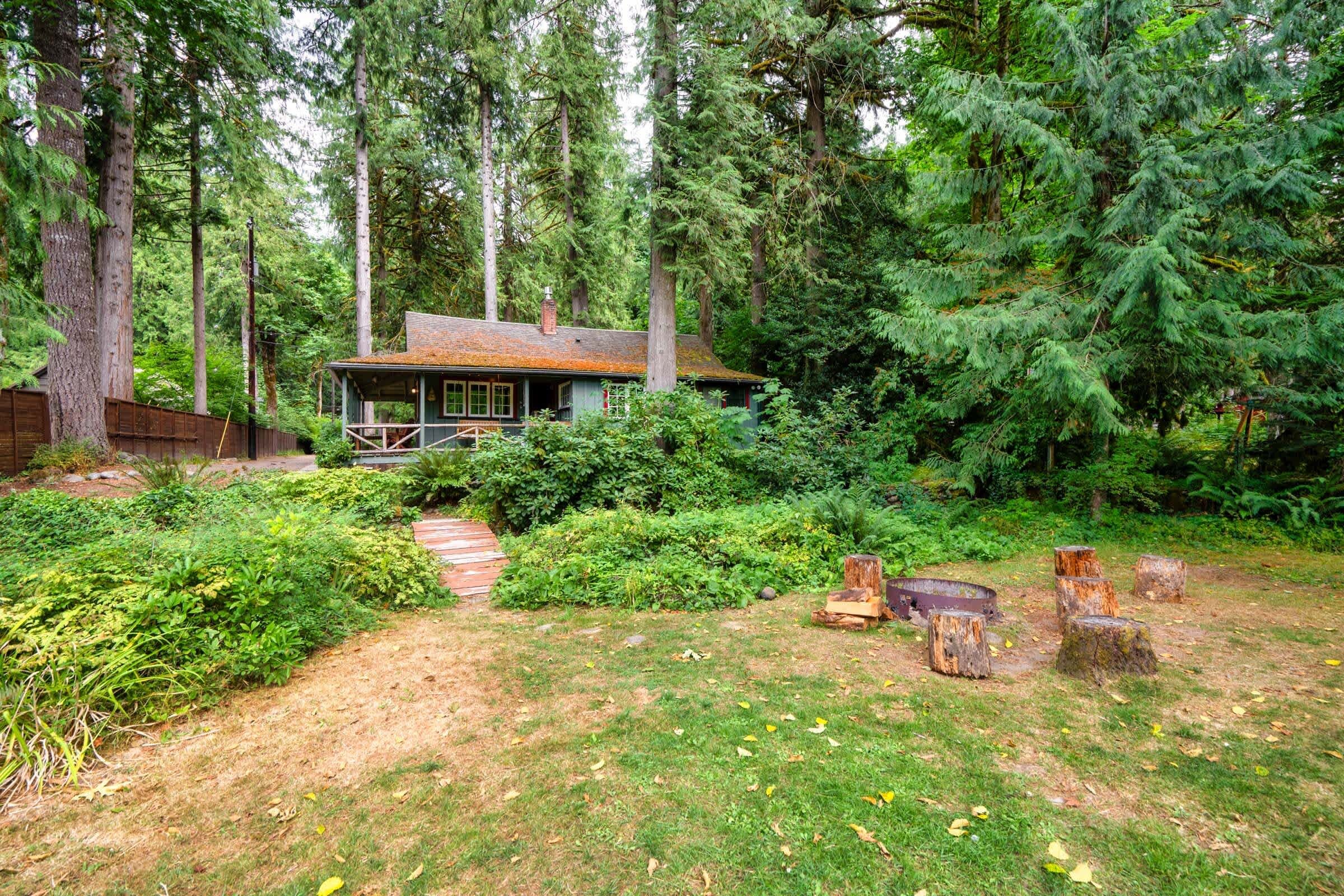 A cabin surrounded by dense forest, with a wooden deck and a pathway leading to the entrance.