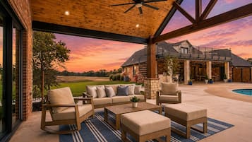 A covered outdoor seating area with a pool, wooden furniture, and a view of a large house.