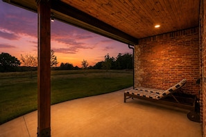A covered outdoor seating area with a bench, a brick wall, and a view of a grassy field during sunset.