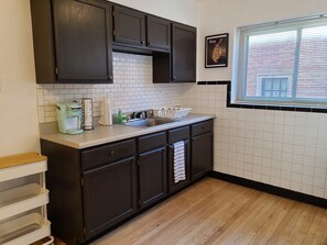 A kitchen with dark cabinets, a white tiled backsplash, a window with blinds, and a countertop with a coffee maker and other items. - A Travel Nurse's Cozy Bird's Nest 1B1B Apartment (St. Louis)