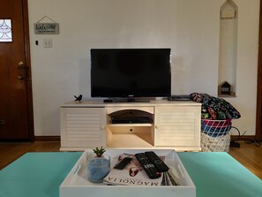 Quarto com televisão sobre suporte, uma pequena estatueta de pássaro, bandeja com controles remotos, vela e revista.