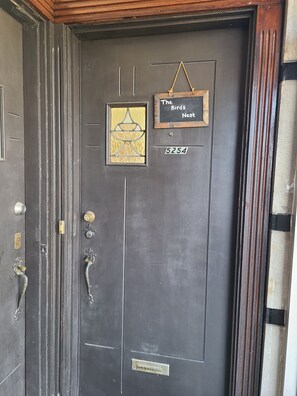 Eine dunkel gefärbte Tür mit einem Messing-Klopfer, ein kleines Fenster mit dekorativem Metallrahmen und ein Schild über der Tür mit der Aufschrift 'The Birds Nest' und der Nummer '5254' darunter.