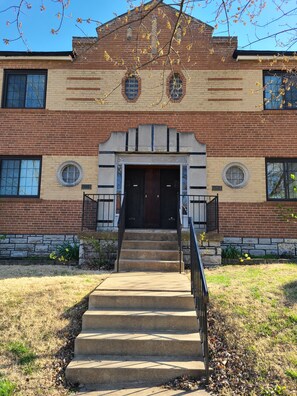 A two-story brick building with a central entrance, a set of concrete steps leading up to the door, and a small garden area with yellow flowers. - A Travel Nurse's Cozy Bird's Nest 1B1B Apartment (St. Louis)