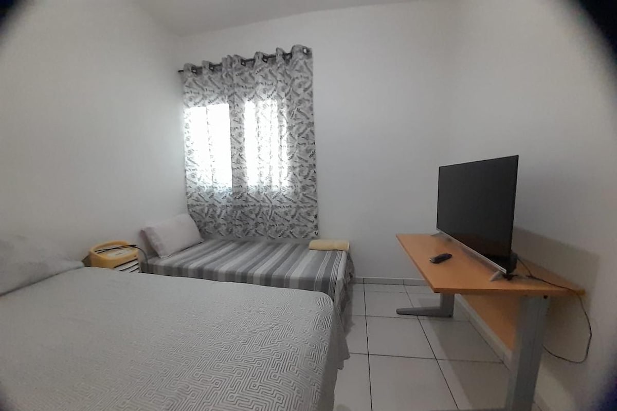 A bedroom with a bed, a desk with a television, and a window with patterned curtains.
