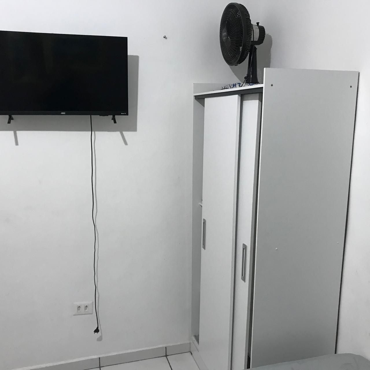 A room with a flat-screen TV mounted on the wall, a white wardrobe with a fan on top, and a visible power outlet on the wall.