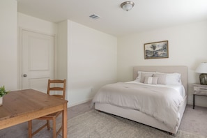 A bedroom with a bed, a nightstand, a lamp, a chair, a table, and a door.
