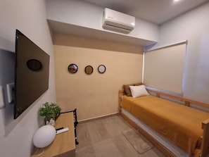 Ein kleines, gemütliches Zimmer mit einem Bett, einem Flachbildfernseher, einem kleinen Tisch mit einer Pflanze und einer Klimaanlage.