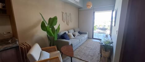 A modern living room with a grey sofa, a wicker chair, a large green plant, and a dining area with a table and chairs.