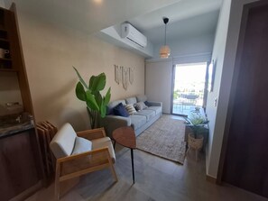 Ein modernes Wohnzimmer mit einem grauen Sofa, einem Korbstuhl, einer großen Grünpflanze und einem Essbereich mit Tisch und Stühlen.