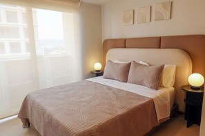 A hotel room with a large bed, two bedside lamps, a window with a view, and wall art. - Zapopan apartment with pool and gym (Zapopan)
