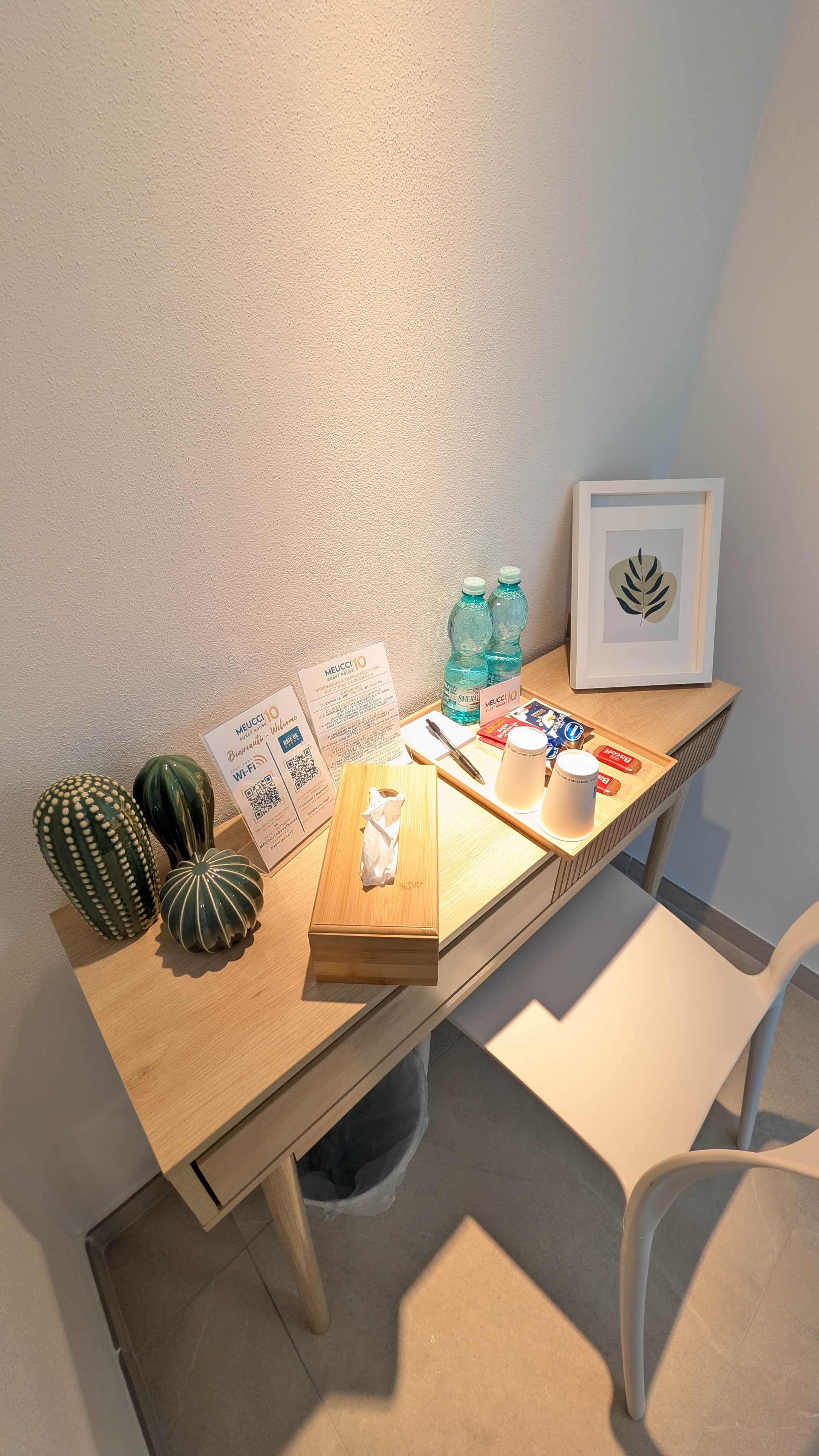 Un escritorio de madera con una planta enmarcada, un calendario, un dispensador de pañuelos, un bolígrafo, una botella de agua y una bandeja con aperitivos.