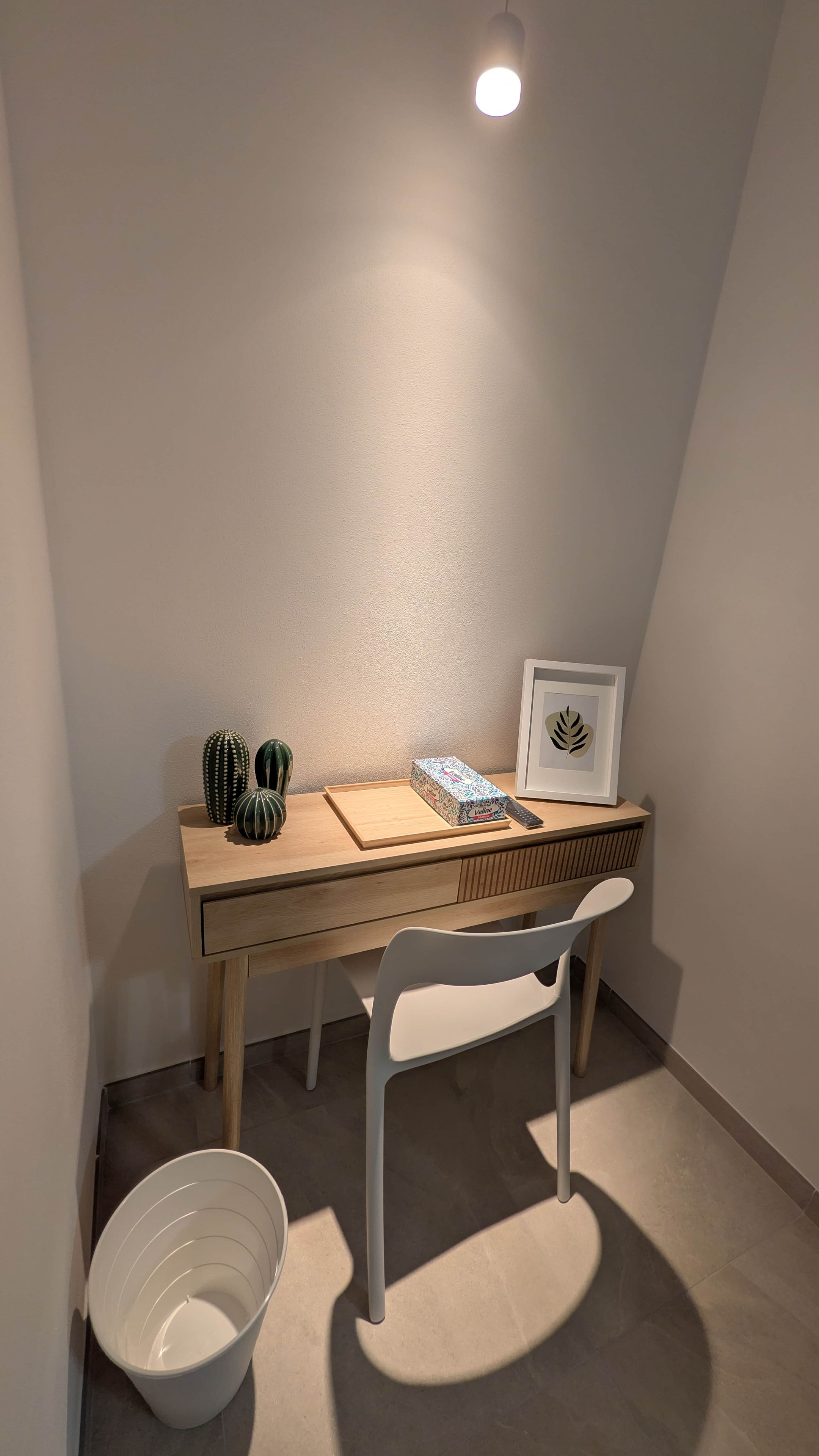 Ein kleiner, gut beleuchteter Raum mit einem Holztisch, einem weißen Stuhl, einem gerahmten Bild und einem Kaktus auf dem Tisch.