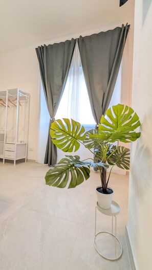 Et værelse med en potteprydet monstera, et hvidt reolsystem og et vindue med grå gardiner.