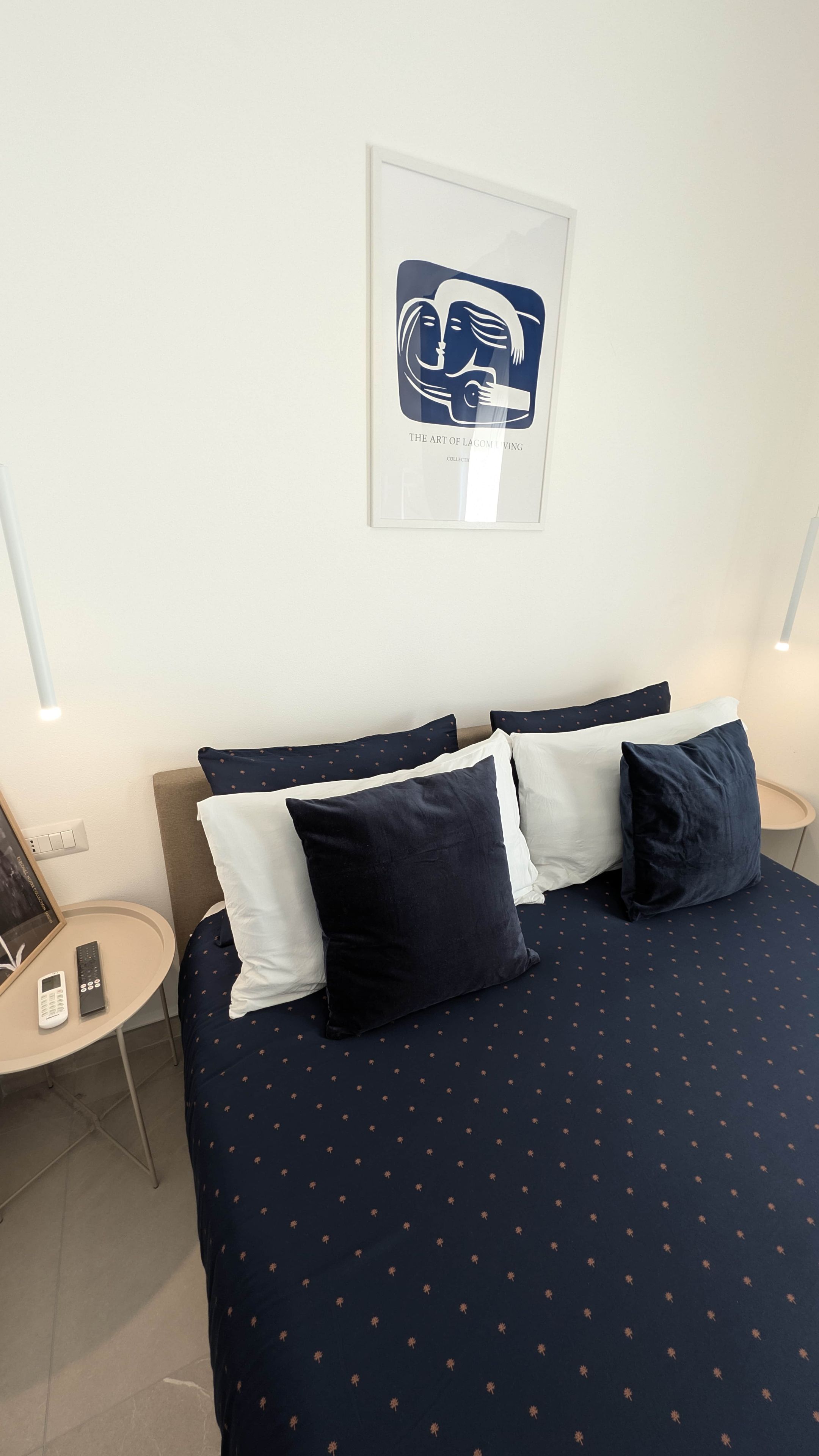 A hotel room with a bed, pillows, a bedside table with a remote control, and a framed artwork on the wall.