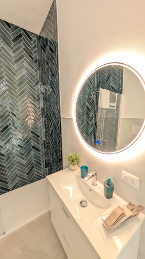 A bathroom with a white sink, a round mirror with a light, and a herringbone-patterned wall. - Molara - Comfort Double Room (Olbia)